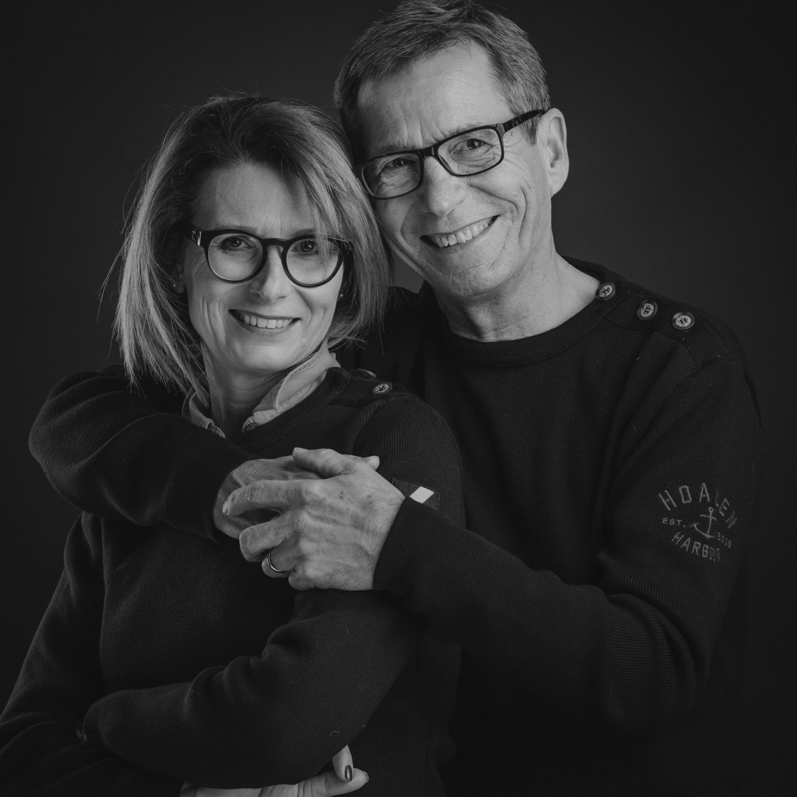 Portrait couple noir et blanc, femme enlacée par son partenaire souriant, lunettes, fond sombre — Photographe couple studio Dol-de-Bretagne, Saint-Malo, Ille-et-Vilaine